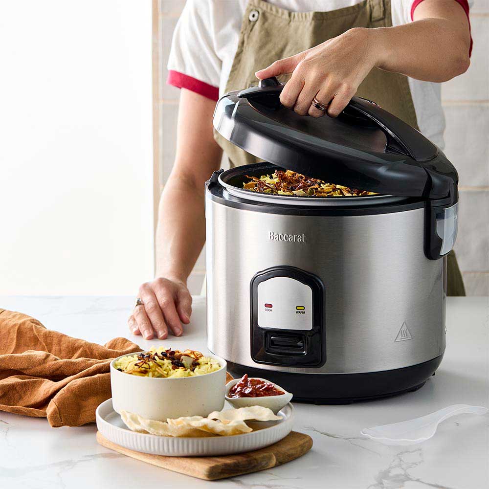 A person opening a silver rice cooker on a kitchen counter next to a bowl of veggies- Baccarat