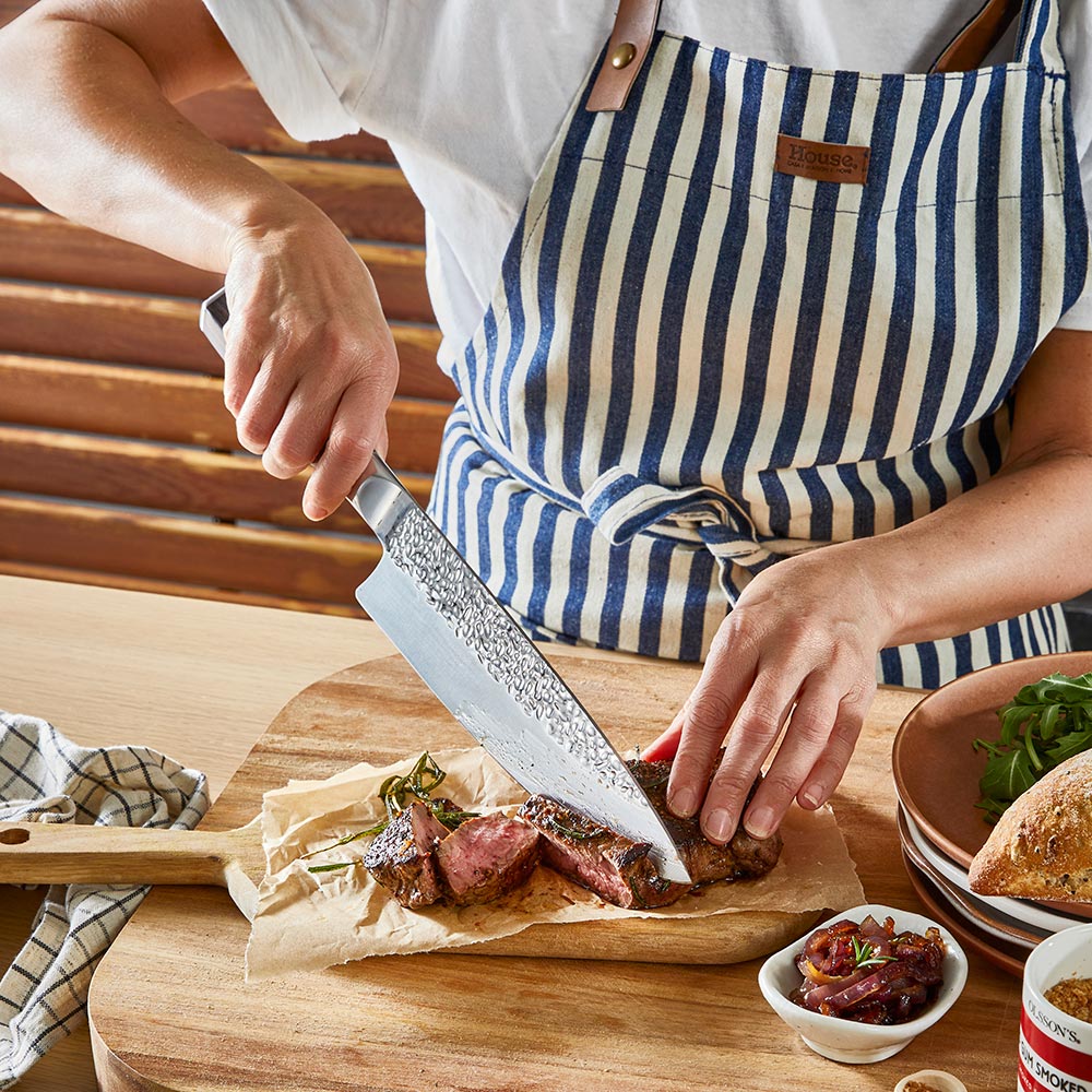 Person slices steak on a wooden board wearing blue-striped apron ideal for kitchen tasks - Baccarat