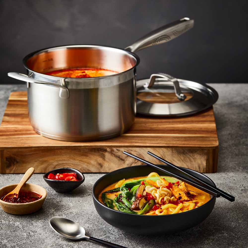 stainless steel saucepan 20cm pot of soup on a wooden board beside black bowl of noodles  - Baccarat