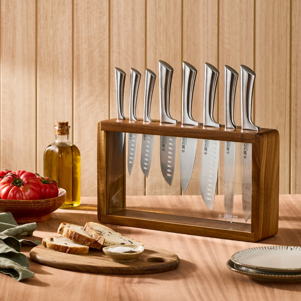 Set of knives in a wooden block on a wooden surface with bread and tomatoes.